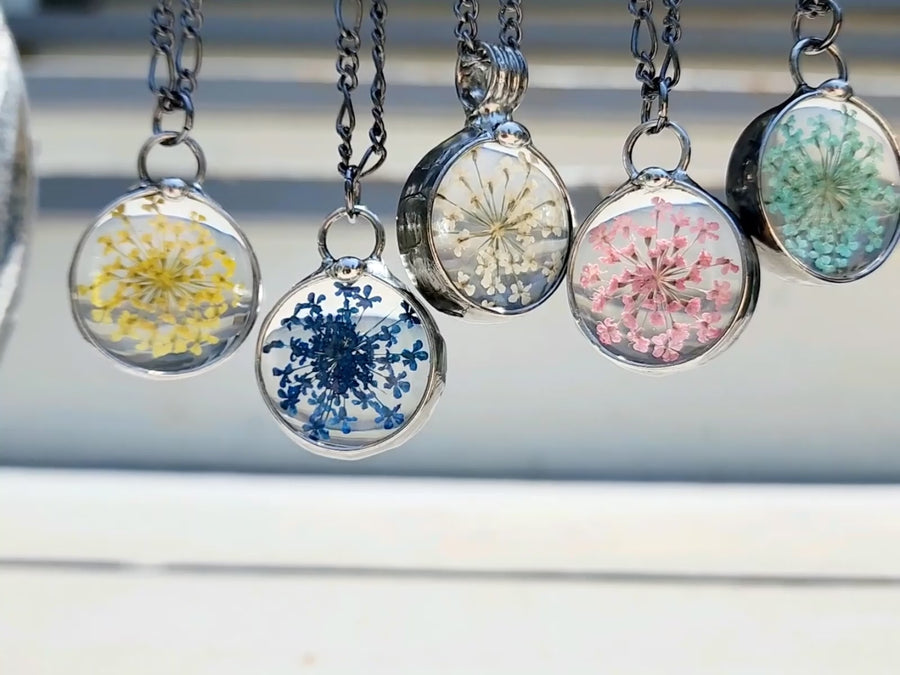 video of 5 queen annes lace pressed flower pendant in assorted colors. Yellow Blue Natural White Pink Aqua. Truly Hand Made in USA by Louisiana Artisan at Bayou Glass Arts Studio. Chain is quality plated fully adjustable Figaro style. All are 100% handmade from metals that contain NO lead, cadmium, zinc or nickel. 