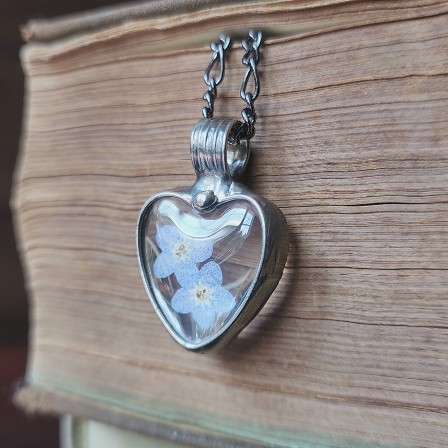Real dry pressed blue flowers, forget me nots, encased under heart shaped glass cabochons form this hand made in USA pendant necklace. Metal work is hand formed with an iron not a torch.