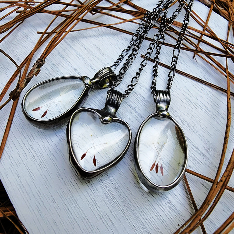 3 dandelion seed pendant necklaces. Left: Teardrop shape with 2 dandelion seeds encased under glass. Center: Heart shaped Pendant with 2 dandelion seeds crossed inside of glass. Right: 3 dandelion Seeds encased in oval glass. All truly hand made in USA by Louisiana Artisan