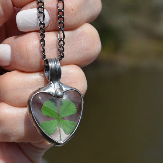 Hand holding a 4 leaf clover glass heart pendant necklace. Truly Hand Made in USA by Louisiana Artisan at Bayou Glass Arts Studio. Chain is quality plated fully adjustable Figaro style. All are 100% handmade from metals that contain NO lead, cadmium, zinc or nickel. 