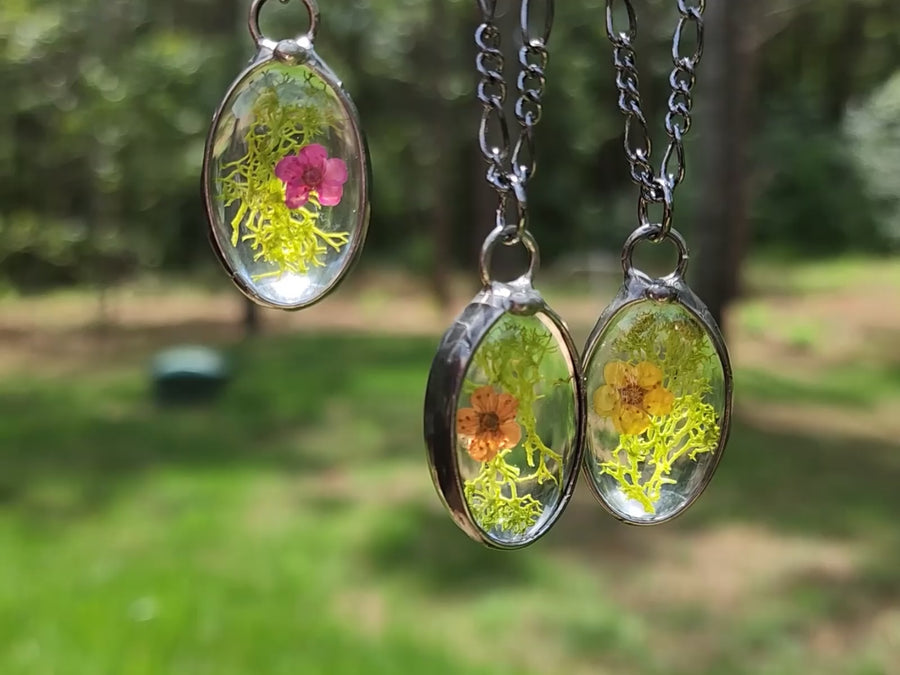 Video of 3 Oval Moss Pendants the first one has a pink floret, the middle one has an orange floret and the one on the right has a yellow floret. The dry pressed florets are dyed forget me not flowers. Handmade in the USA.