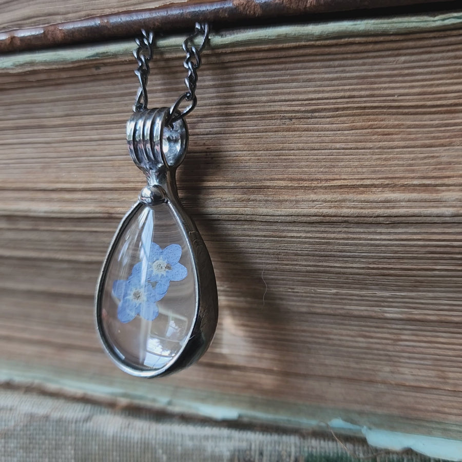 Real dry pressed blue flowers, forget me nots, encased in teardrop shaped glass cabochons form this hand made in USA pendant necklace. Metal work is hand formed with an iron not a torch.