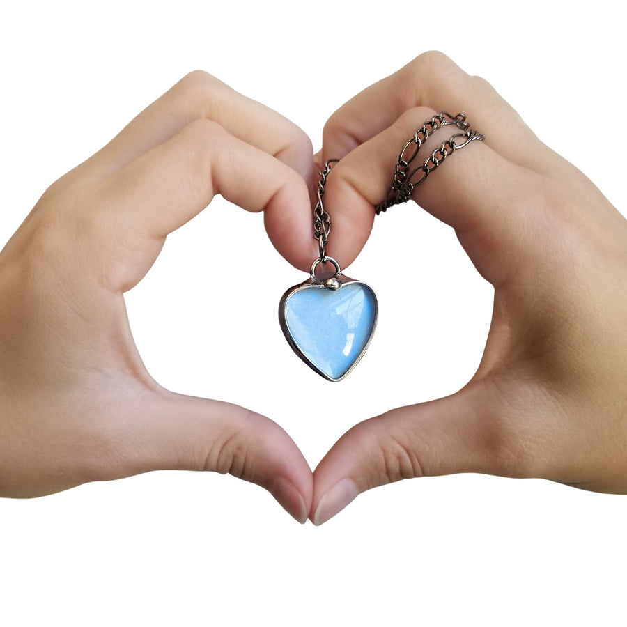Heart Hands holding a Blue See See Heart Pendant Necklace with white background. See See Jewelry is hand made in USA by Louisiana Artisan at Bayou Glass Arts Studio. Secret message hidden inside is revealed when the pendant is held up to a light.