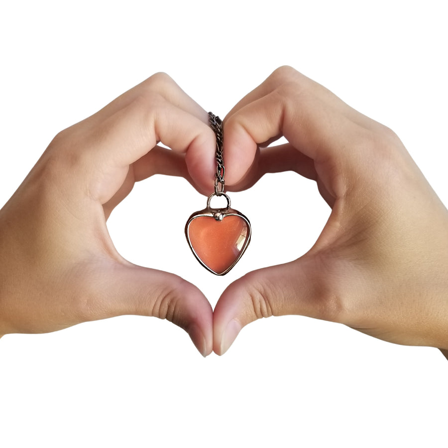 Handmade See See Orange Heart Pendant Necklace wrapped in heart hands with white background. See See Jewelry is hand made in USA by Louisiana Artisan at Bayou Glass Arts Studio. Secret message hidden inside when pendant is held up to a light. 